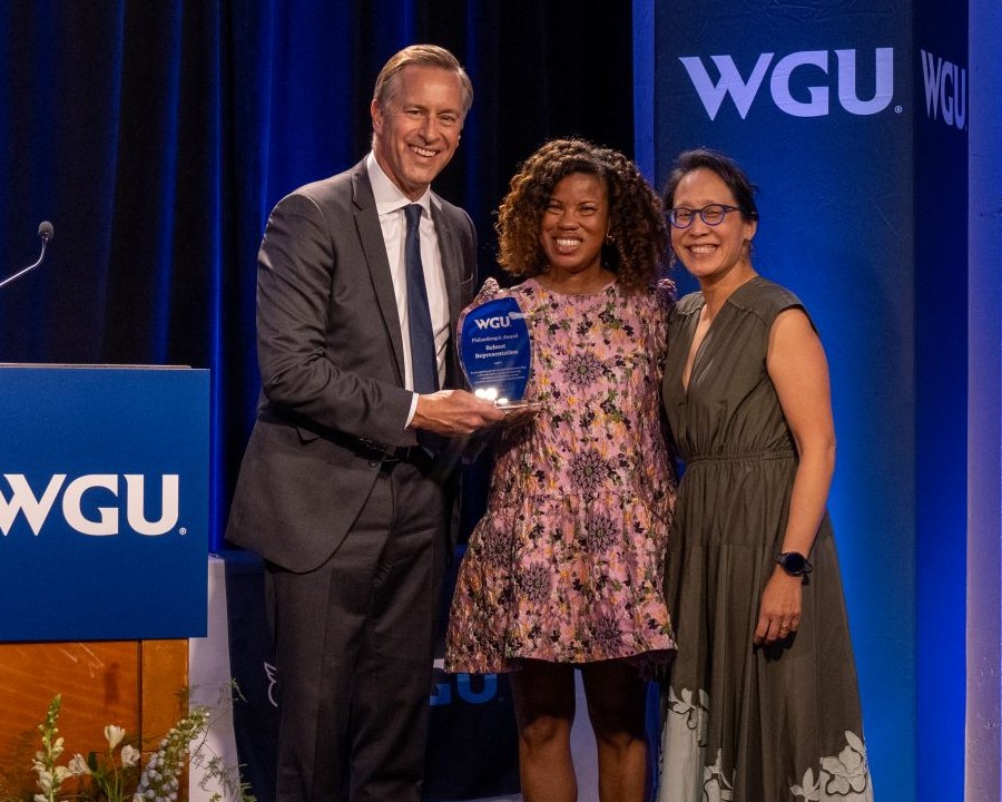 Three smiling leaders pose onstage at a WGU event as a man presents a crystal recognition award to a woman.