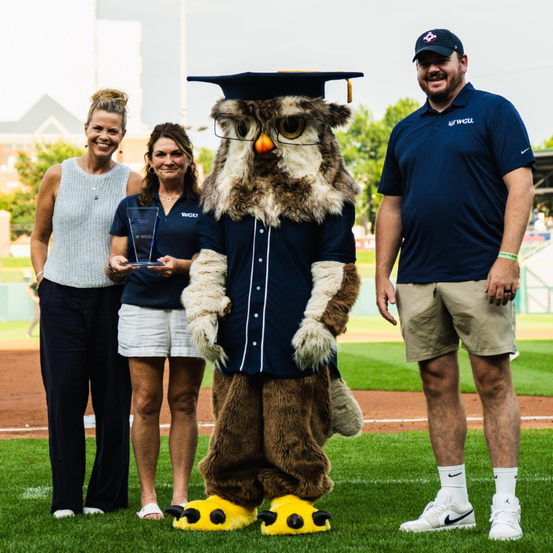 A graduation-capped owl mascot stands with three WGU staff members on the field at a baseball stadium.
