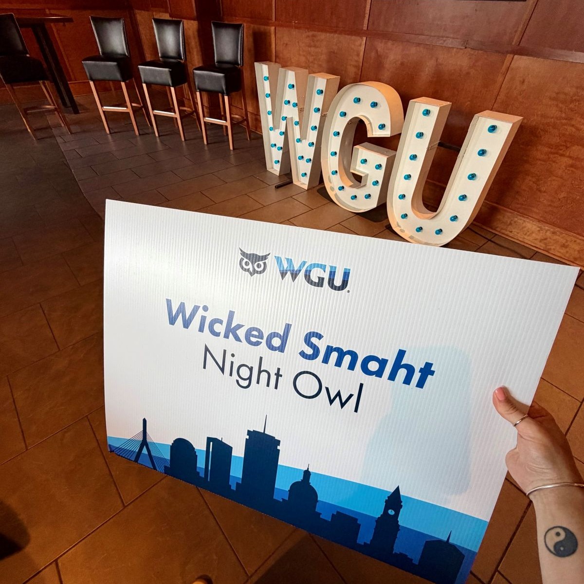WGU Wicked Smaht Night Owl sign held before illuminated WGU letters and bar stools at an indoor event.