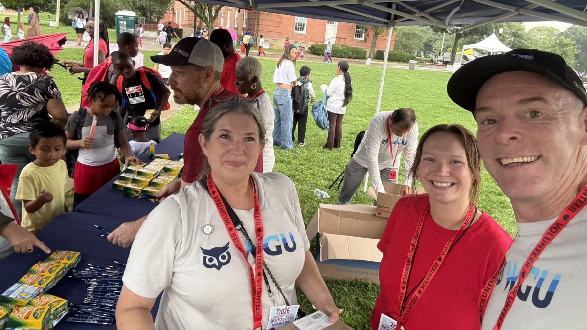 Three Volunteers in white and red shirts staff a community giveaway table as families gather at an outdoor event.