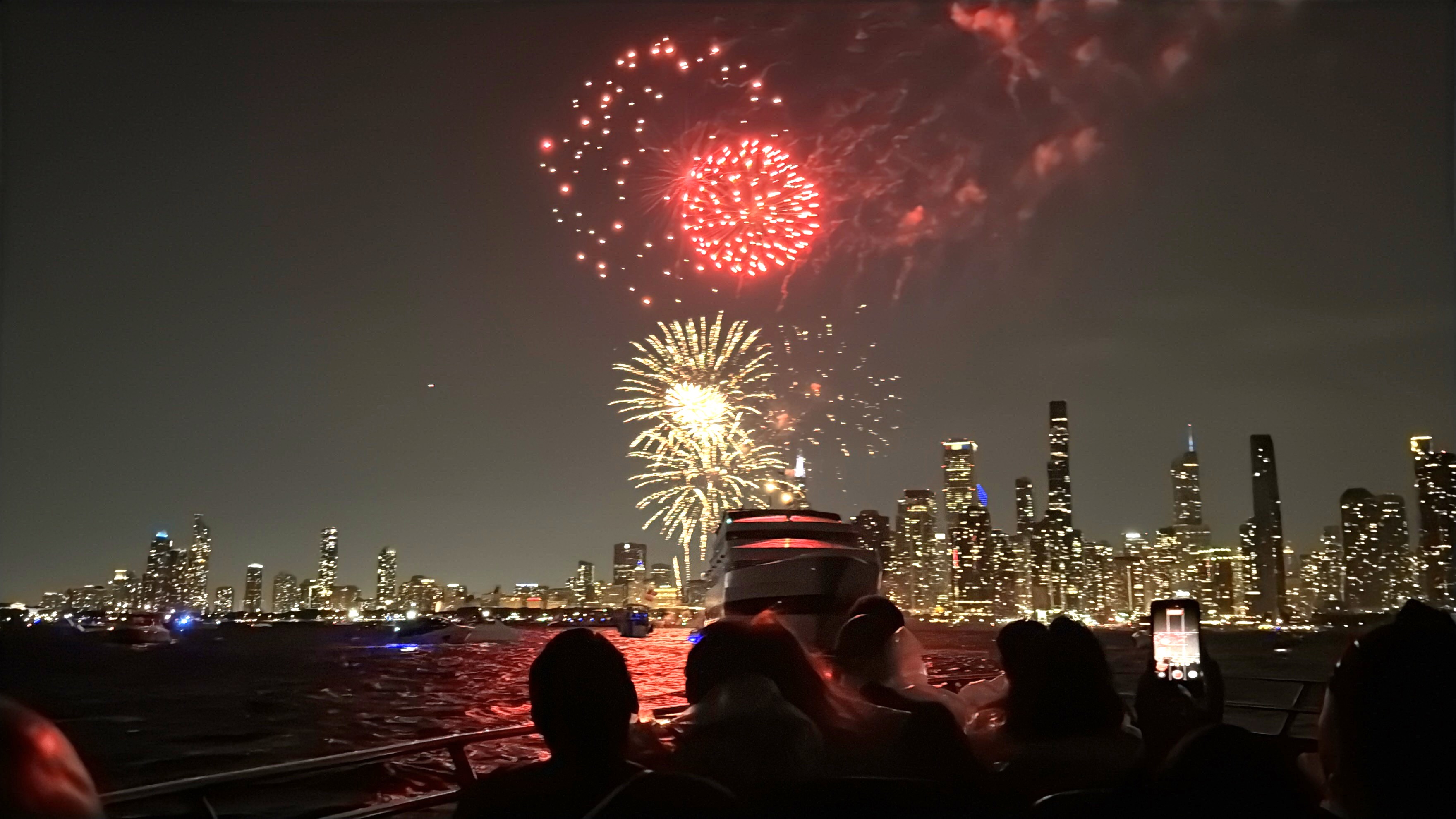 Red and gold fireworks burst above Chicago's illuminated skyline as passengers watch from a boat at night.