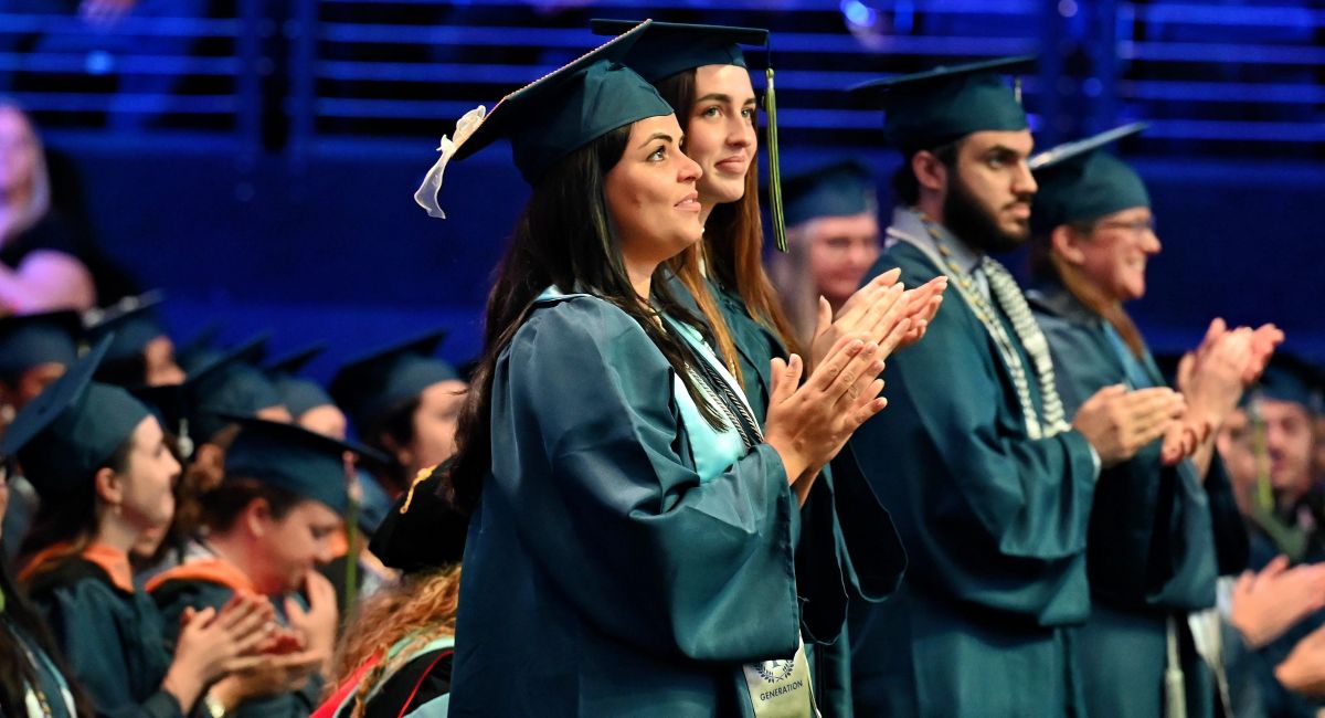Graduates in blue caps and gowns stand and applaud during a ceremony in a blue-lit auditorium filled with fellow students.
