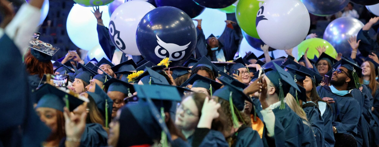 A large group of graduates in caps and gowns celebrating with colorful balloons, some featuring an owl logo.