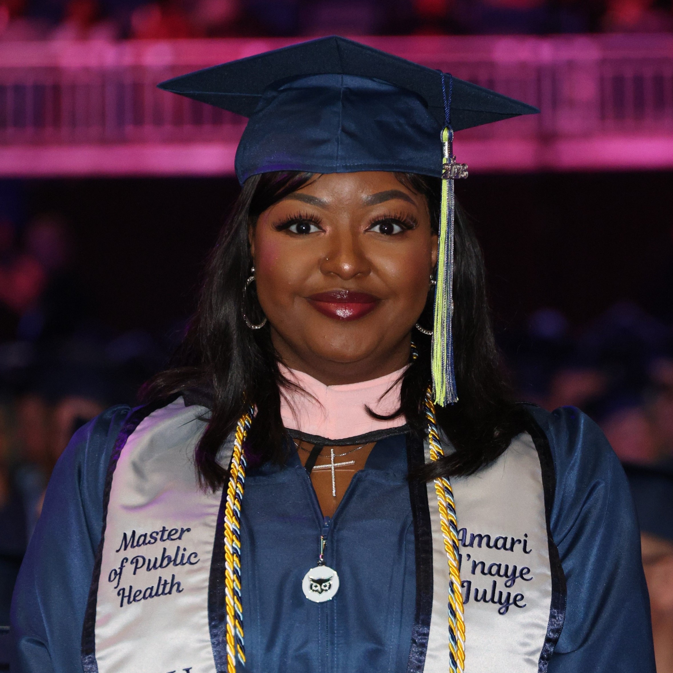 A smiling graduate in a navy blue cap and gown with a stole that reads "Master of Public Health"