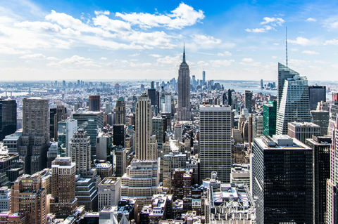 Panoramic view of the New York City skyline, featuring the Empire State Building and numerous skyscrapers under a bright blue sky with white clouds.