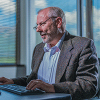 A man wearing glasses sits at a desk, typing on a computer keyboard