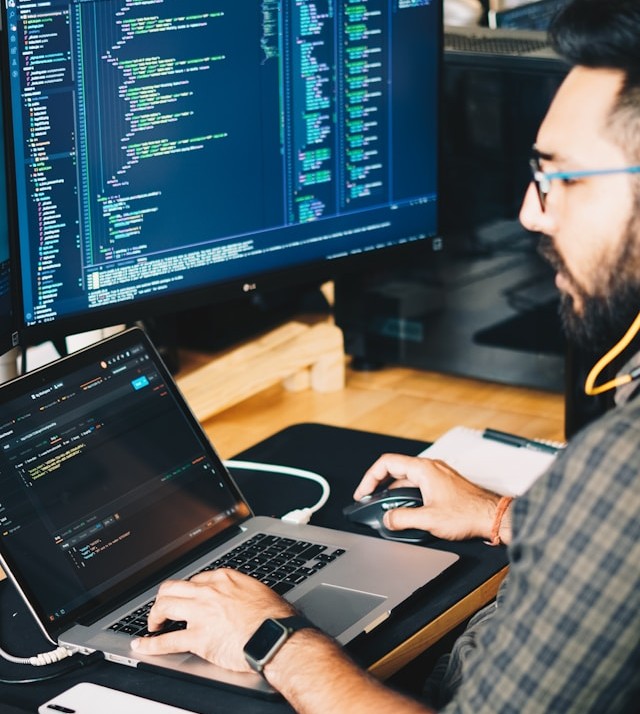 A developer intently coding on a laptop and an external monitor displaying lines of code.