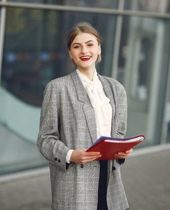 Smiling businesswoman in a gray plaid blazer holds a red folder outside a modern glass office building.