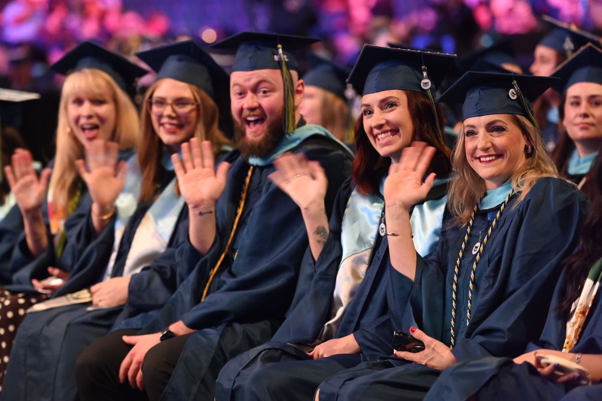 A group of happy graduates in navy caps and gowns smiling and waving during a ceremony.