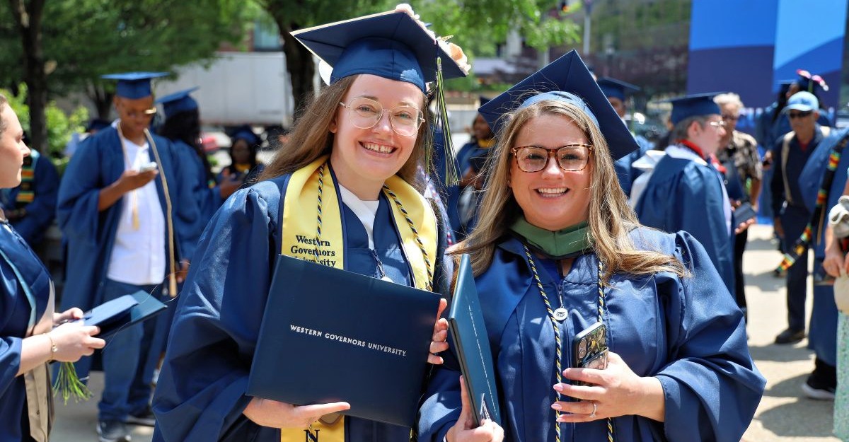 Two smiling women in blue graduation gowns and caps hold diplomas, celebrating their achievement.