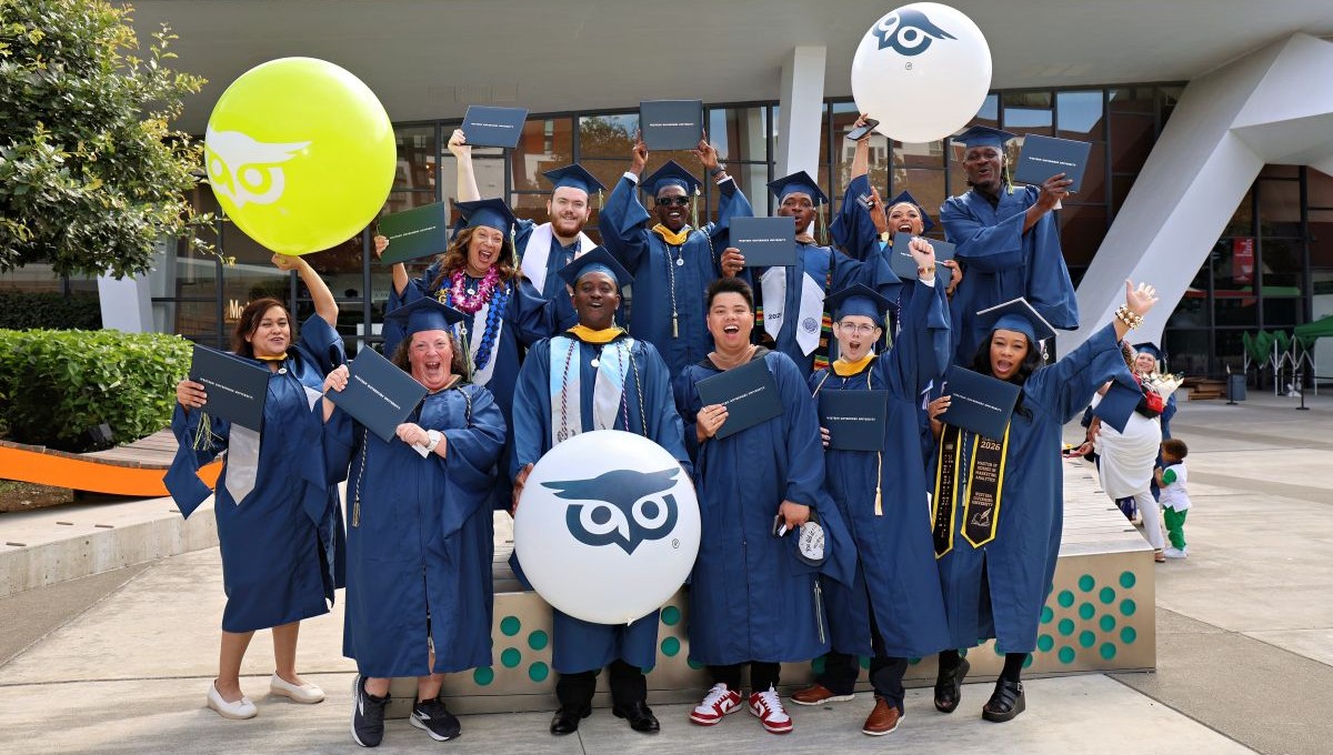 A jubilant group of graduates in blue caps and gowns hold diplomas and pose with large owl-logo balloons outdoors.