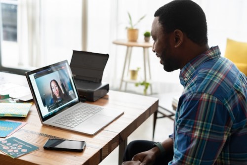 A man in a plaid shirt smiles while participating in a video call with a woman on his laptop.