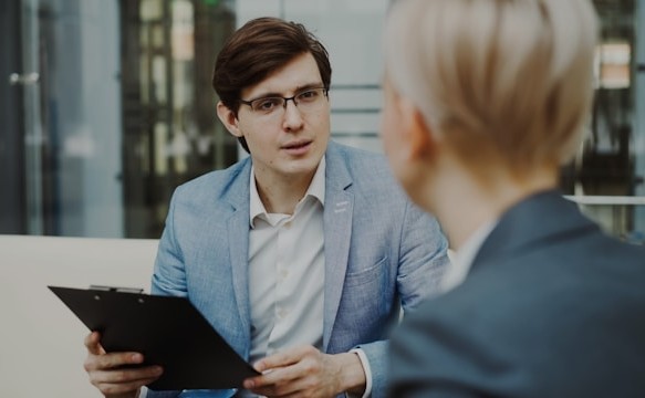 Business professional holding a clipboard while speaking with a colleague in a modern office setting.