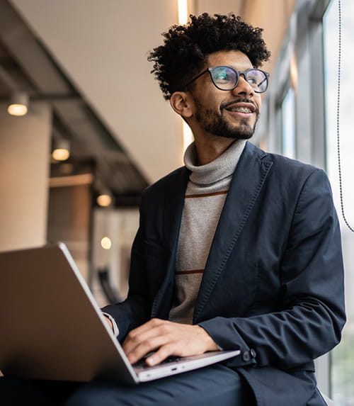 Smiling professional in glasses works on a laptop by a window, looking outward in a modern office setting.