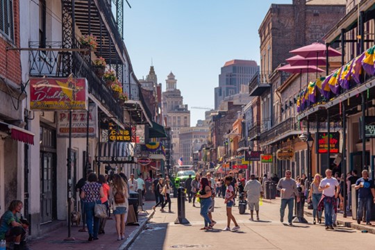 A bustling street scene in New Orleans, with people walking along a street lined with multi-story buildings featuring balconies and colorful awnings.