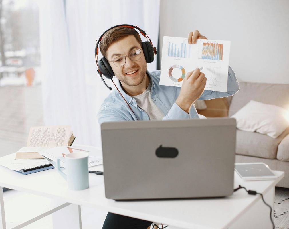 A man wearing headphones and glasses points to a document with charts and graphs while on a video call in front of his laptop.