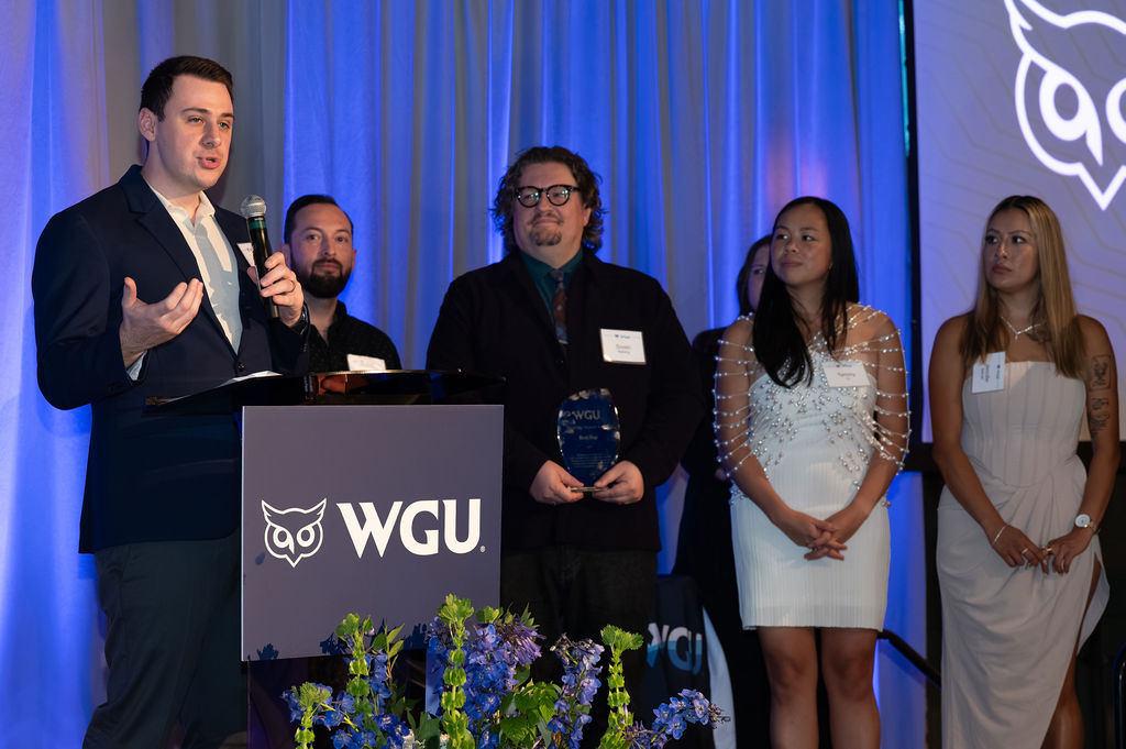 A speaker addresses attendees at a WGU awards ceremony as an honoree holds a glass trophy beside fellow guests.