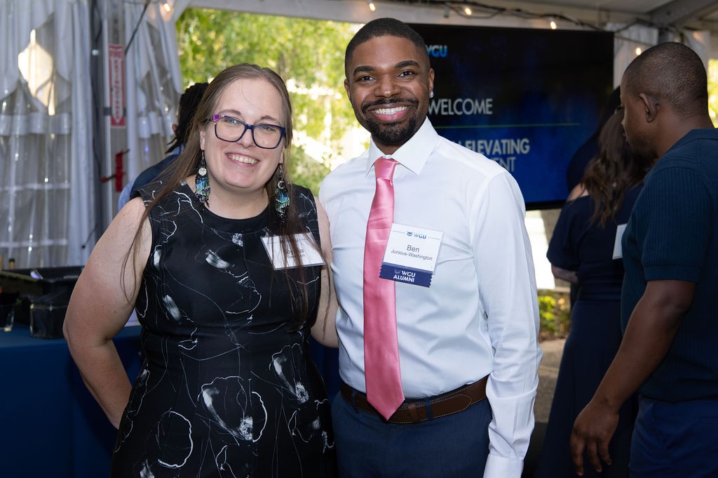 Two smiling WGU alumni wearing name badges pose together at a welcome event beneath a tent with string lights.