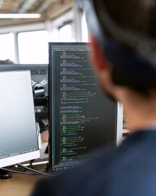 Developer viewed from behind works at a dual-monitor desk, with lines of colorful code displayed on one screen.