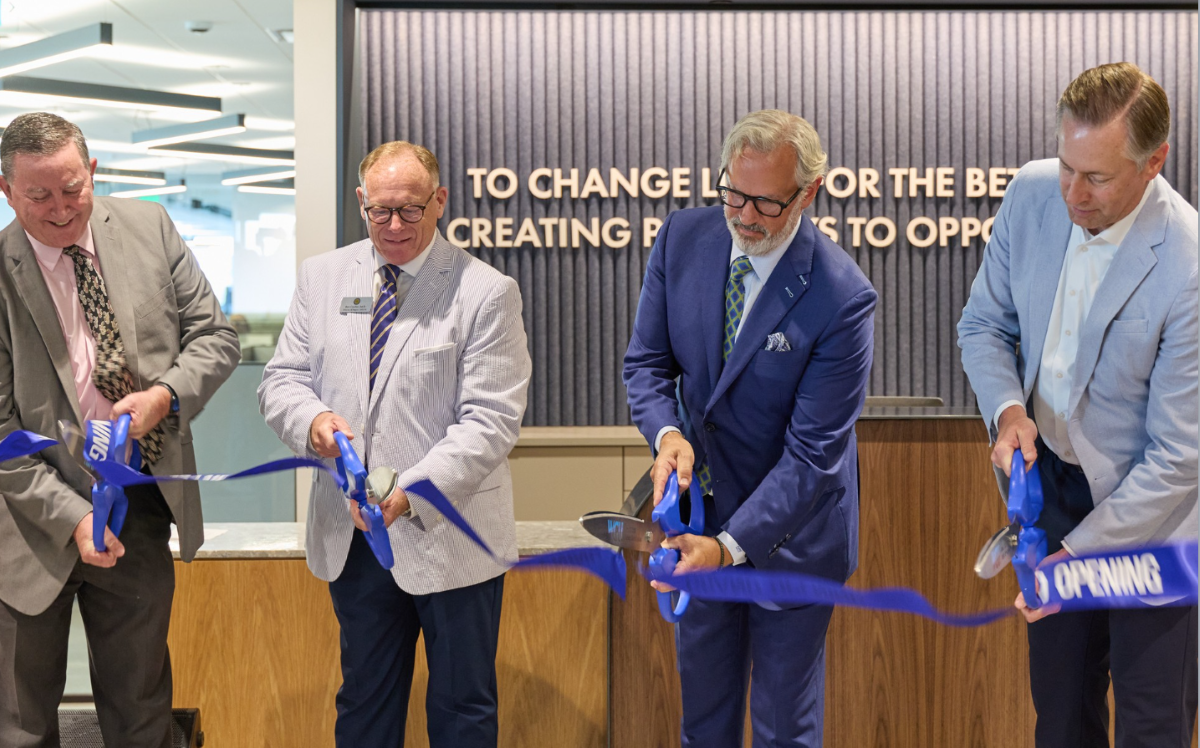 Four executives cut a blue opening ribbon with ceremonial scissors together during a modern office launch event.