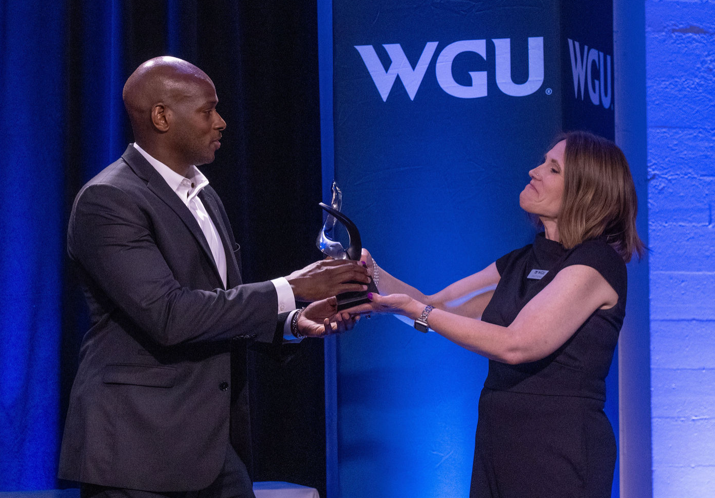 A man in a suit accepts a glass award from a woman onstage beside a Western Governors University banner.