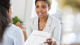 A professional Black woman speaks while handing a document and pen to another person.