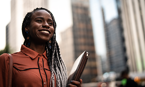 A smiling Black woman with dreadlocks in an orange shirt holds a portfolio while standing in a city.