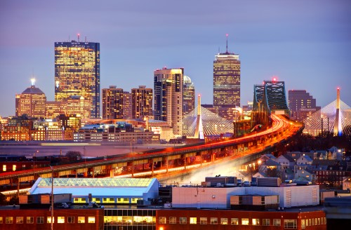 Panoramic view of Boston at dusk, featuring bright buildings and a multi-lane bridge with traffic lights.