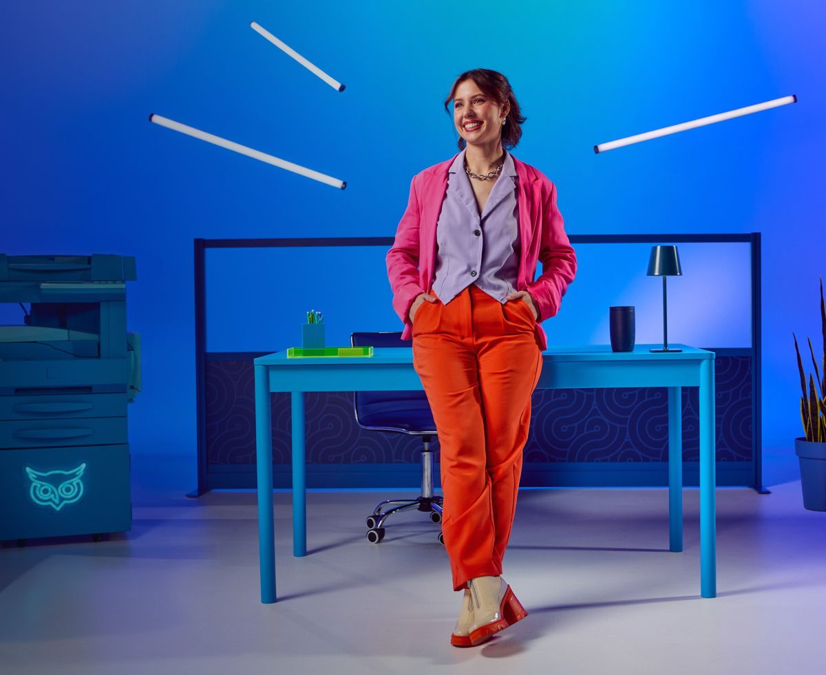 A smiling woman in a pink blazer and orange pants leans casually against a blue desk in a vibrant blue office setting with floating neon lights.