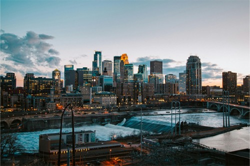 Twilight view of Minneapolis skyline above the Mississippi River, with bridges, traffic, and a low waterfall.